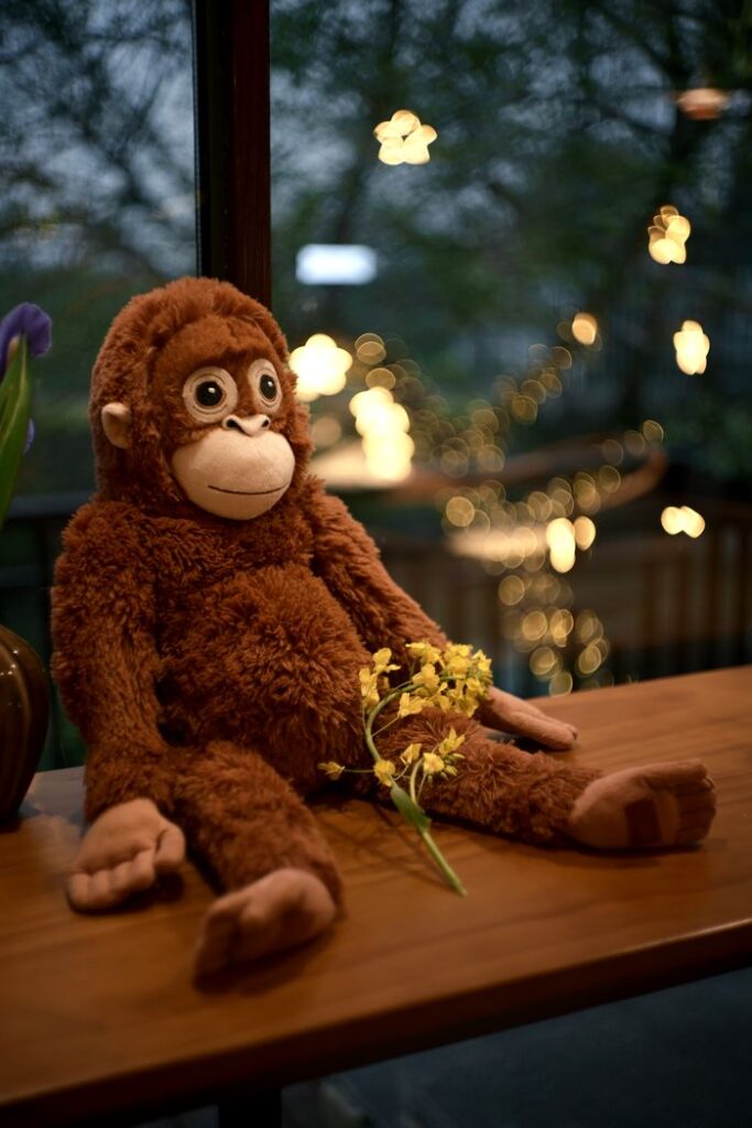 A charming brown stuffed monkey toy sits on a wooden table with a small bouquet of yellow flowers.
